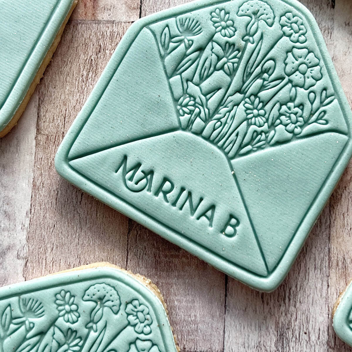 Green cookie stamp with floral patterns and 'Marina B' branding on a wooden surface.