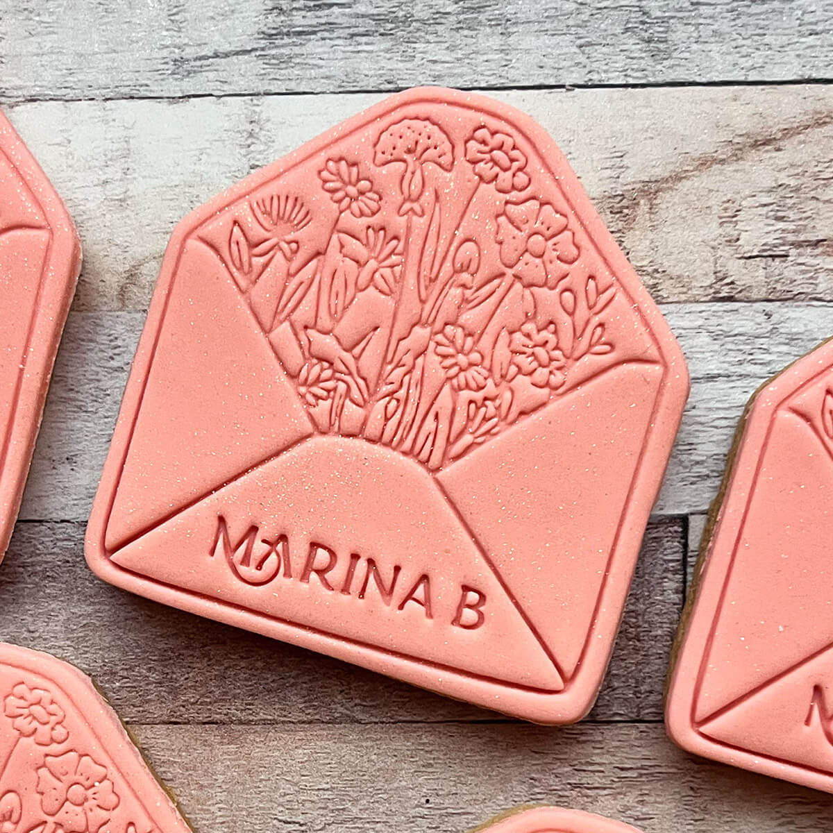Pink cookie with floral design and 'Marina B' branding on a wooden surface