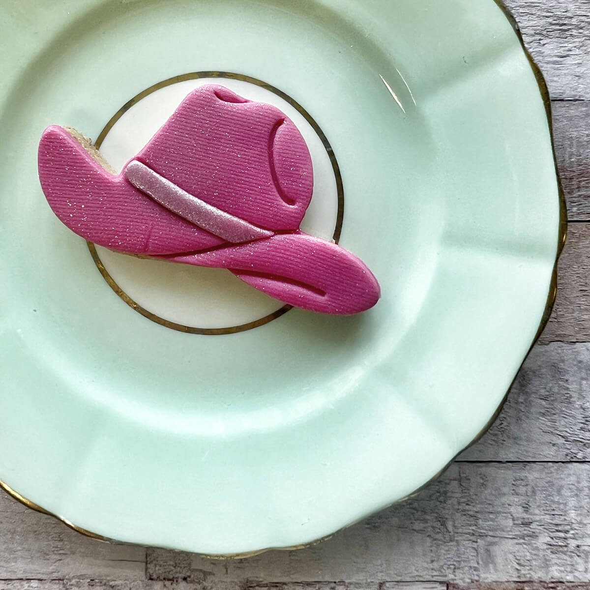 Pink cowboy hat cookie on a light green plate with gold rim on a wooden surface
